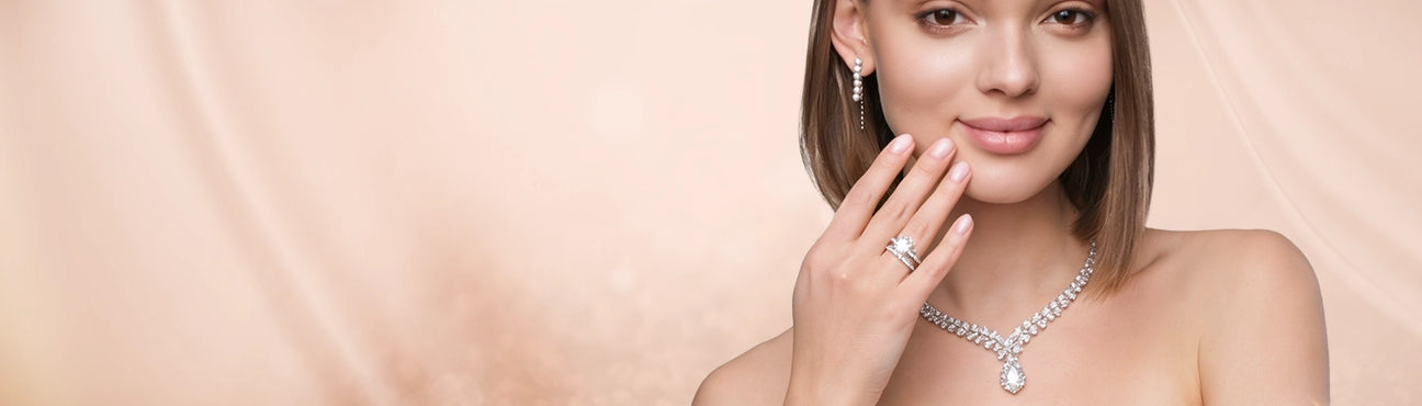 A woman models a sparkling lab grown diamond necklace and matching earrings and engagement ring against a soft beige background